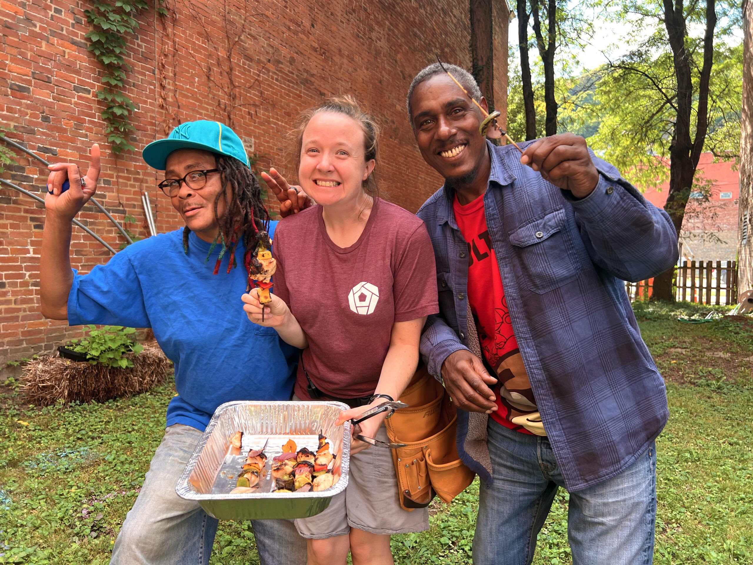 Three people standing together in a garden with smiles on their faces and holding up grilled chicken and vegetable kabobs.